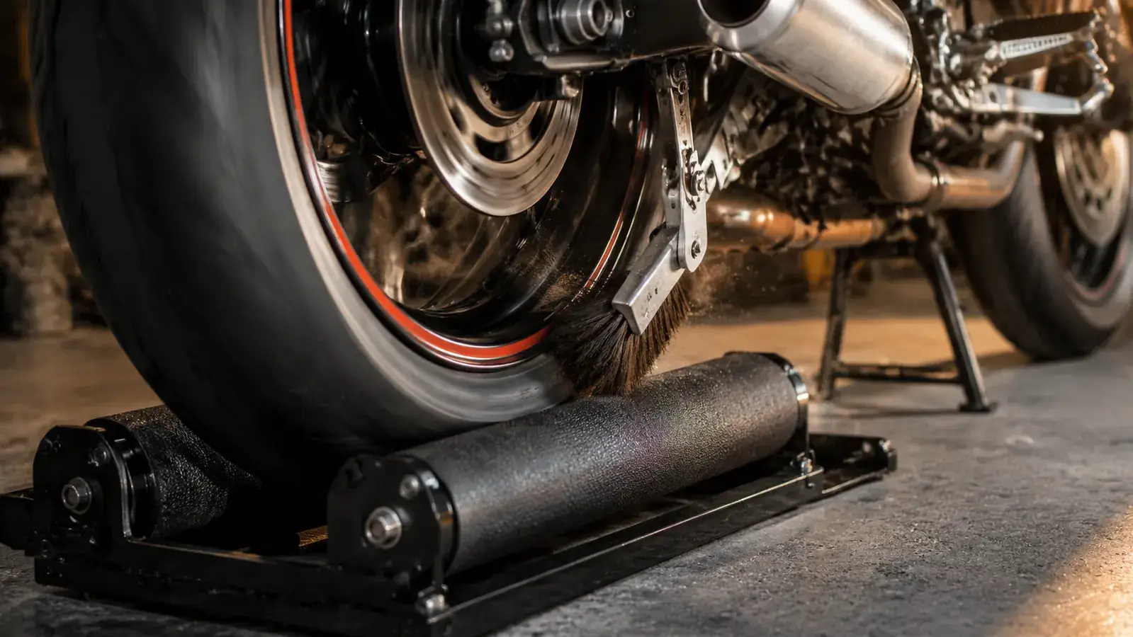 Close view of the rear wheel spinning freely on the two padded roller arms of the stand while a brush cleans the tire sidewall