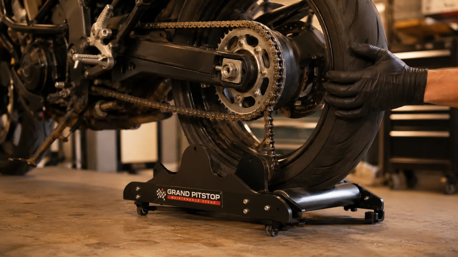 A gloved hand turning the rear wheel by hand while it spins on the roller stand, the chain slack around the sprocket ready to be cleaned