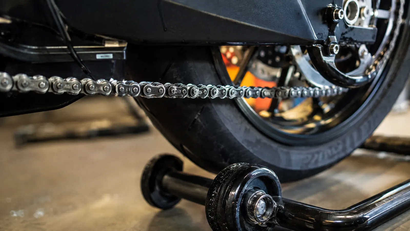 A close-up of a clean motorcycle chain with a drop of fresh lube on the rollers, rear wheel raised off the floor
